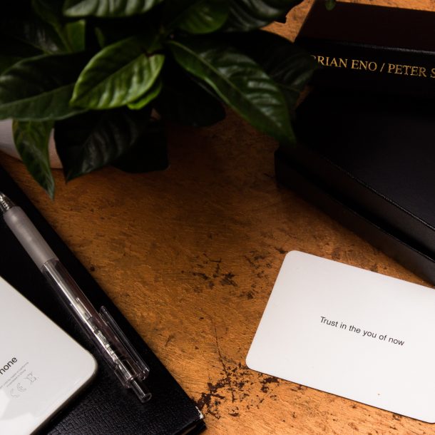 A white card that says "Trust in the you of now" sits on a brown table next to to black boxes, an iphone, and a plant. The card is from a pack designed to help people get inspiration, and possible grow your business.