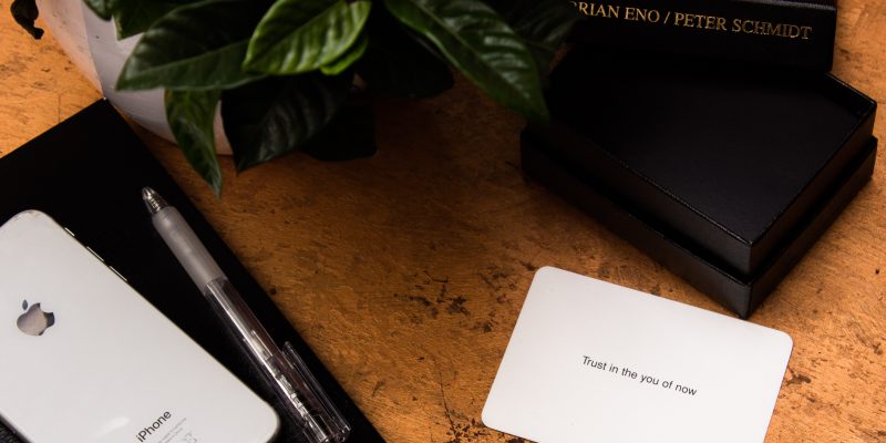 A white card that says "Trust in the you of now" sits on a brown table next to to black boxes, an iphone, and a plant. The card is from a pack designed to help people get inspiration, and possible grow your business.