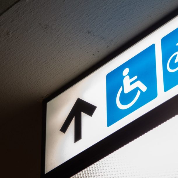 A fluorescent white sign on which there is a blue image of a person sitting in a wheelchair. Next to the image is a black arrow pointing forward. The image as a whole accompanies an article about increasing accessibility in one's business.