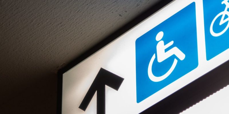 A fluorescent white sign on which there is a blue image of a person sitting in a wheelchair. Next to the image is a black arrow pointing forward. The image as a whole accompanies an article about increasing accessibility in one's business.
