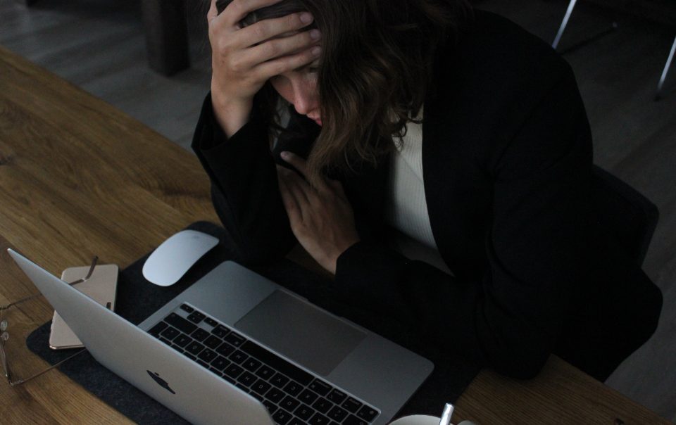 A woman sitting at a desk in front of a laptop with her head in her hands. It accompanies an article about hustle culture.
