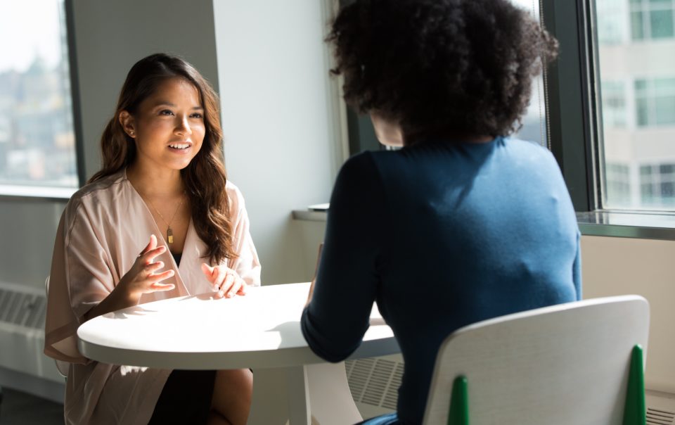 Two people sitting at a round table. One has long brown hair and is facing the viewer, and the other has an afro and is facing away from the viewer. The photo accompanies an article about business coaching.